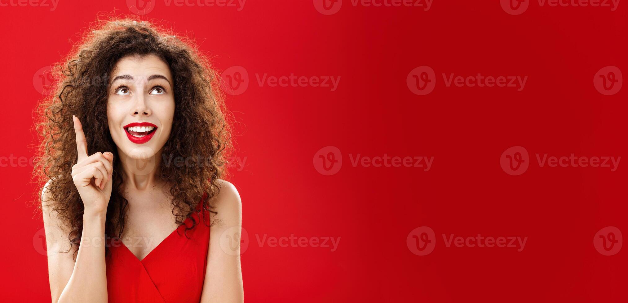 Portrait of delighted charming silly female with curly hairstyle in dress raising index finger in eureka gesture smiling happily looking up having great plan or idea posing charismatic over red wall photo
