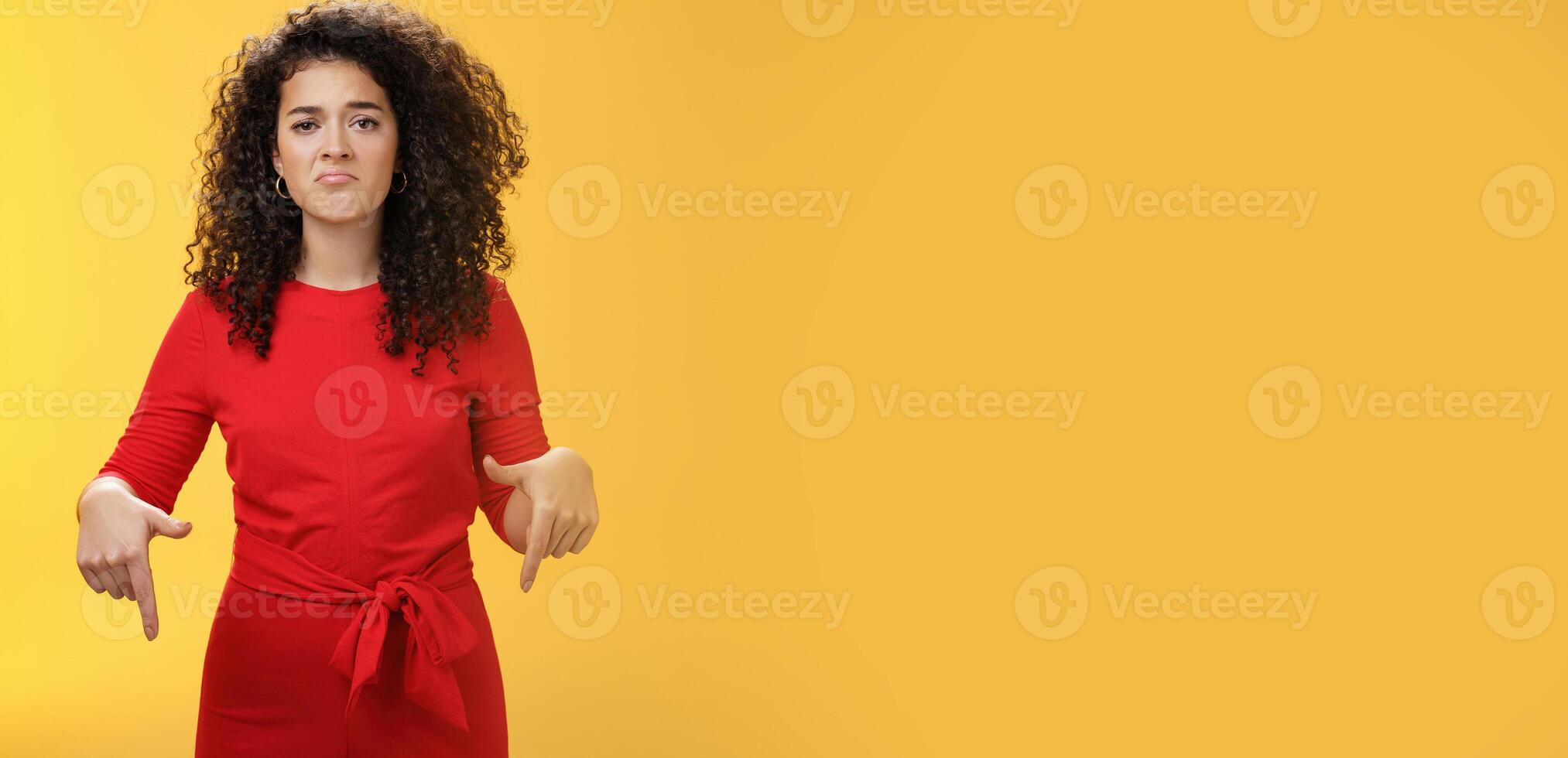 Portrait of gloomy girl with sad face pursing lips down frowning disappointed and pointing down with regret and unsatisfied expression, missing opportunity feeling upset over yellow background photo