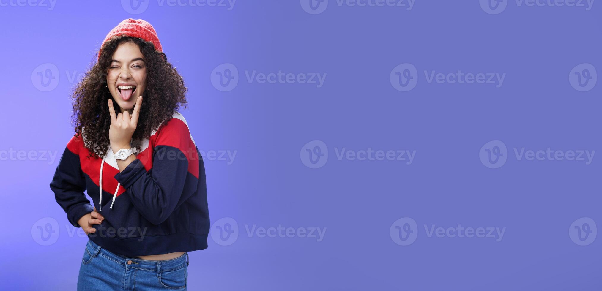Stylish and cool energized swag girl with curly hairstyle in warm beanie showing rock-n-roll gesture near mouth and sticking out tongue smiling having fun, partying and enjoying life over blue wall photo