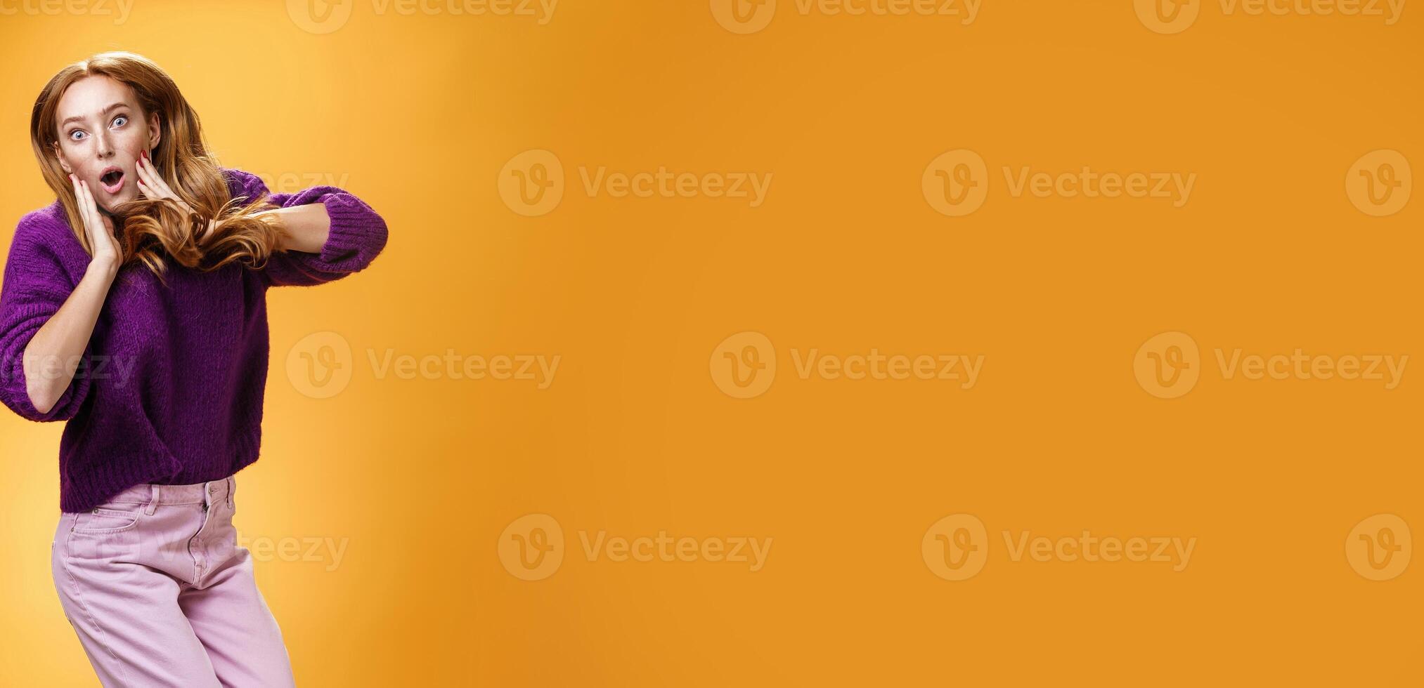 Impressed and surprised amazed good-looking redhead female in purple sweater open mouth astonished and holding hands on cheeks as jumping popping eyes amused over orange background photo