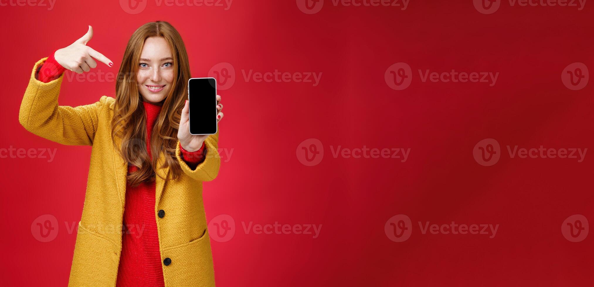 Pick this phone you never regret. Portrait of friendly-looking attractive and confident redhead glamour girlfriend in yellow coat showing smartphone pointing at mobile as smiling broadly at camera photo