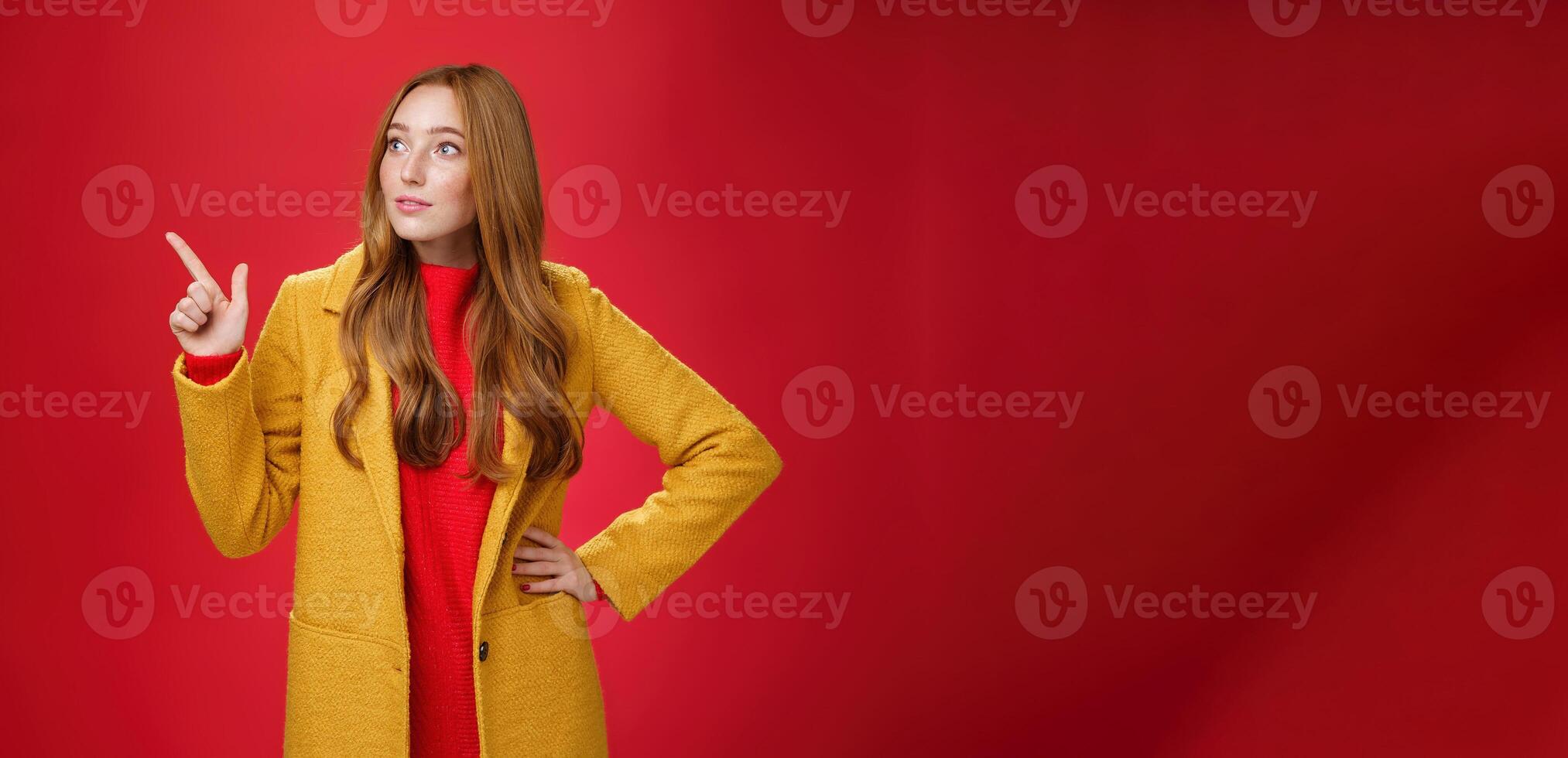 Wondered curious good-looking redhead woman in coat stopping on way looking and pointing at upper left corner curious and interested, observing surprising promotion over red background photo