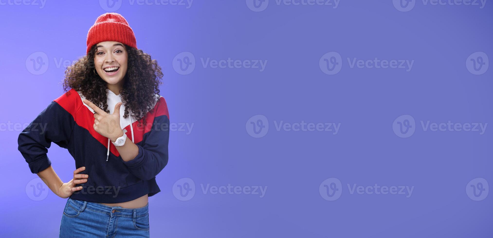 Stylish and sociable friendly-looking attractive woman with curly hair in warm beanie pointing at upper left corner laughing from joy and amazement having fun posing happily over blue background photo