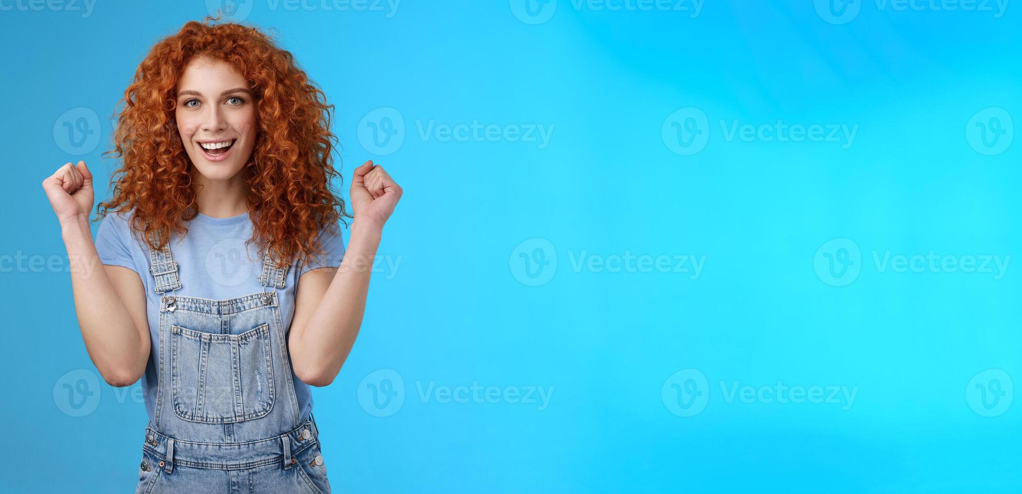 Hooray successful day. Cheerful chrismastic redhead girl summer dungarees clench fists joyfully smiling broadly determined achieve success, triumphing winning game standing upbeat blue background photo