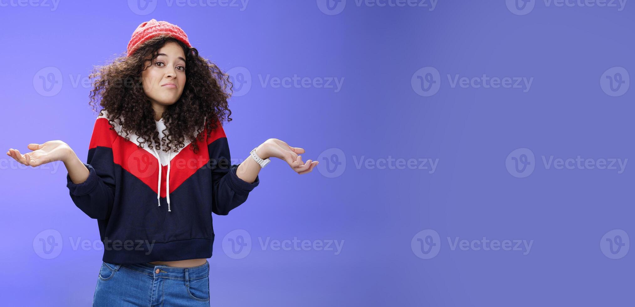 Upset girl cannot get clue what happened shrugging turning away as pouting feeling questioned and unsure what answer, standing clueless and puzzled in sweatshirt and stylish hat over blue background photo