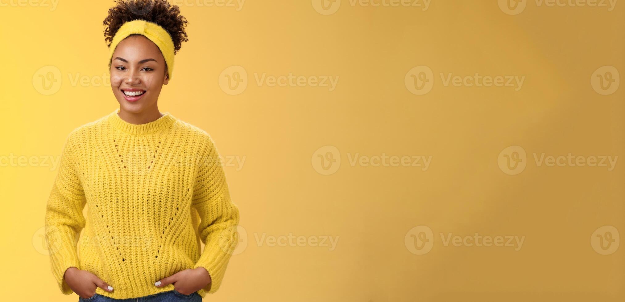 Friendly outgoing relaxed young african-american beautiful girl headband forehead in sweater hold hands pockets smiling broadly have fun talking interesting communication, standing yellow wall photo