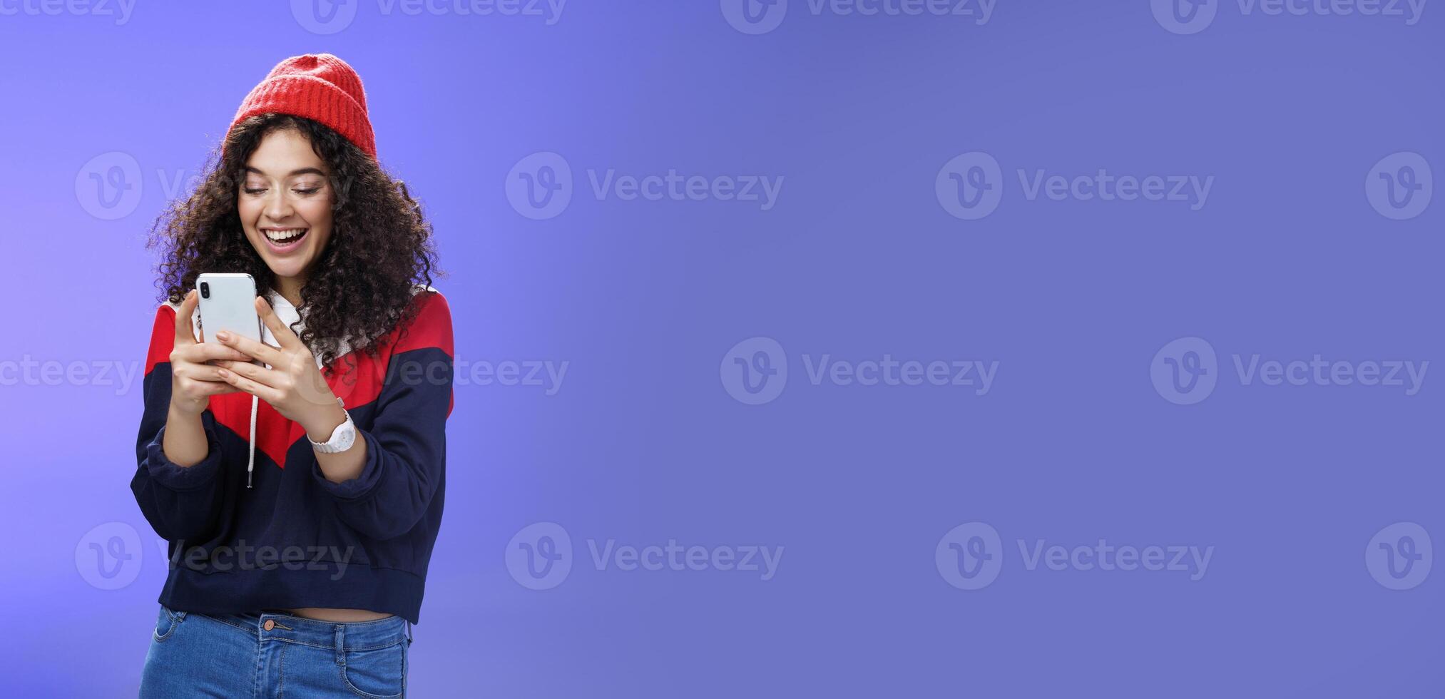 Joyful and sociable stylish caucasian woman with curls in warm beanie having fun laughing and chuckling as using smartphone typing funny message looking at mobile phone screen happily over blue wall photo