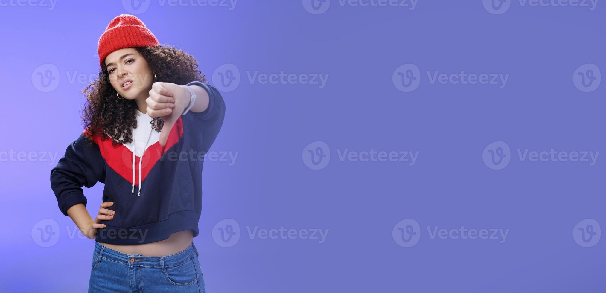 Nah dislike. Portrait of annoyed and unimpressed arrogant cool girl with curly hair tilting head with distain and scorn showing thumbs down in disappointment grimacing, posing over blue wall photo