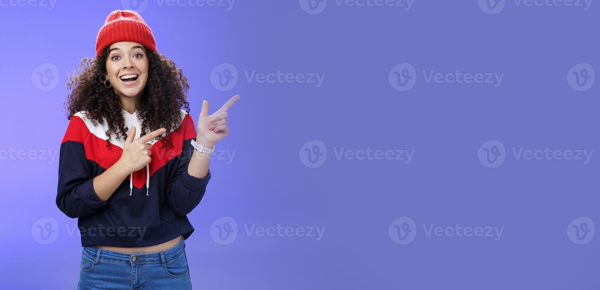 Enthusiastic friendly-looking attractive young 20s woman with curly hair in warm beanie and sweatshirt smiling open mouth amazed and delighted as pointing at upper right corner over blue background photo