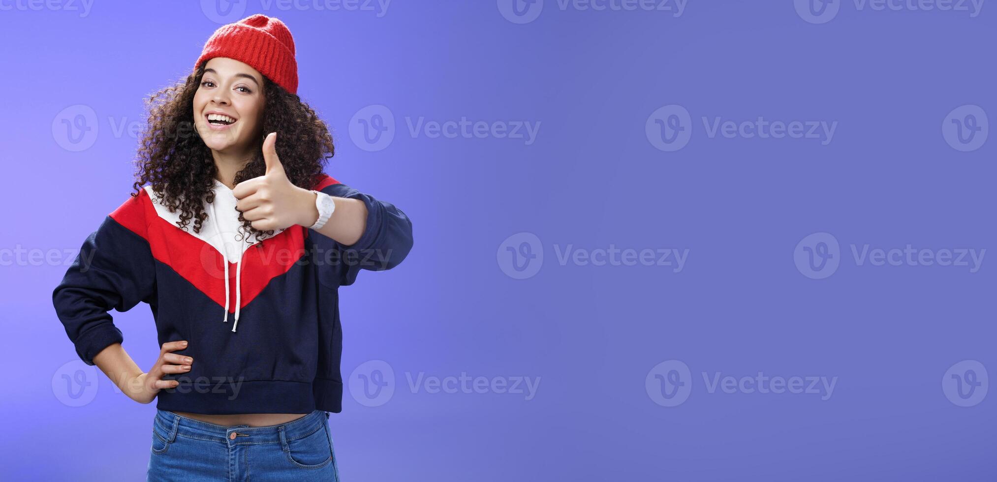Nice work, keep up. Delighted and supportive charming sportswoman in winter beanie, sweatshirt and watch showing thumbs up with satisfied smile, giving approval, liking and cheering great effort photo