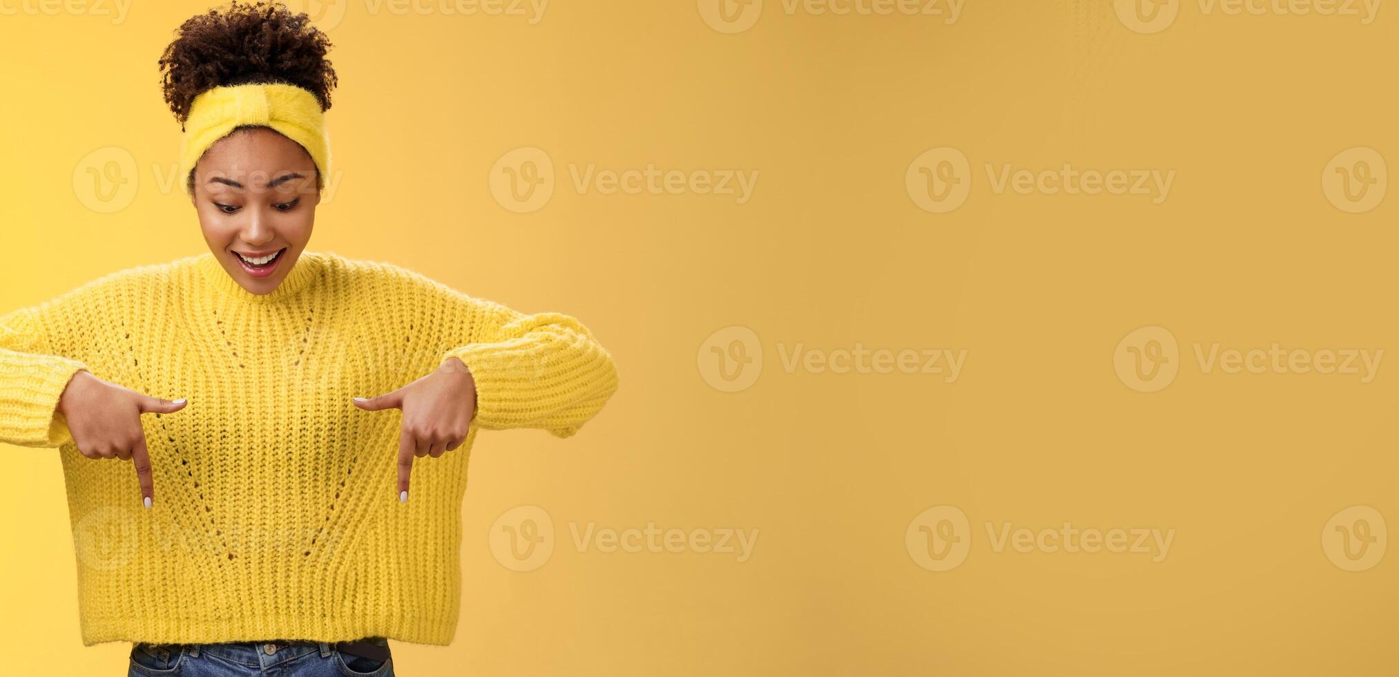 Impressed enthusiastic smiling fascianted african-american woman in sweater headband look pointing down index fingers gasping astonished fascinated cool thrilling promo, standing yellow background photo
