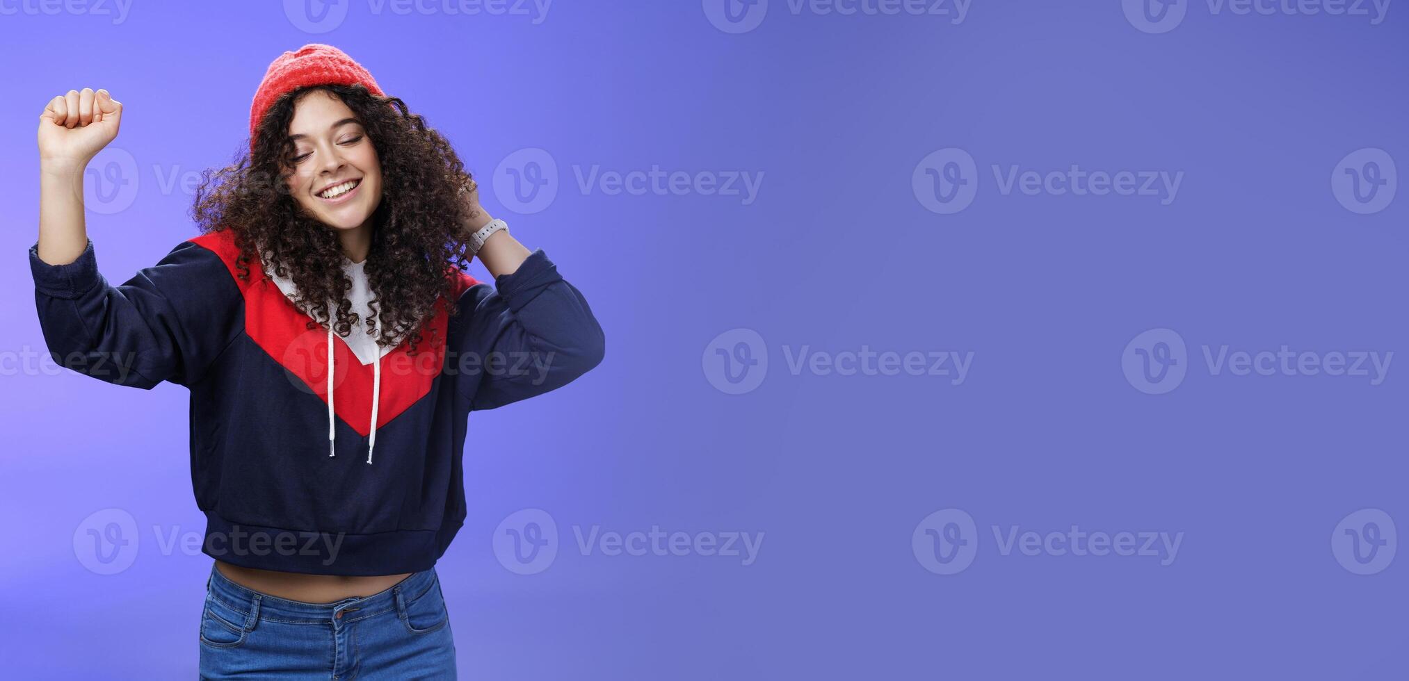 Girl delighted and carefree dancing over blue background with closed eyes and tender smile moving raised hands in rhythm of music, dreaming having positive mood wearing warm winter hat and sweatshirt photo