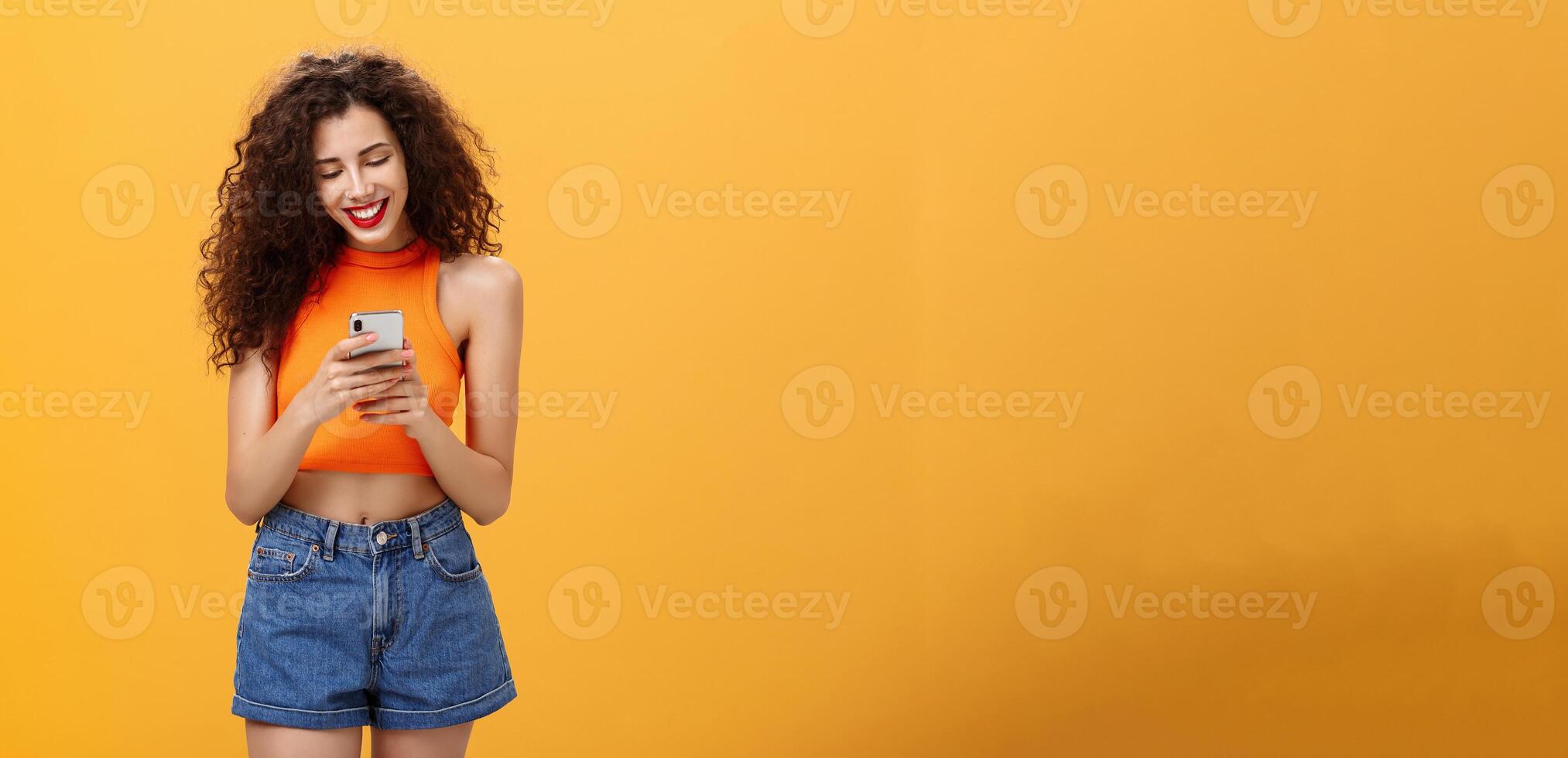 Girl spending time in internet texting friend messages via smartphone laughing while looking at device screen standing happy and upbeat over orange background in cropped top and denim shorts photo