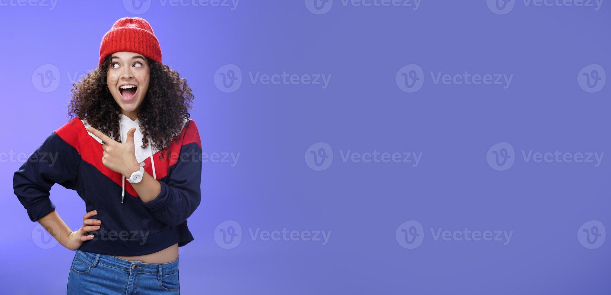 Cute and playful girl in winter stylish outfit and watch smiling broadly with excitement and joy pointing, looking at upper left corner thrilled and joyful as posing over blue background photo
