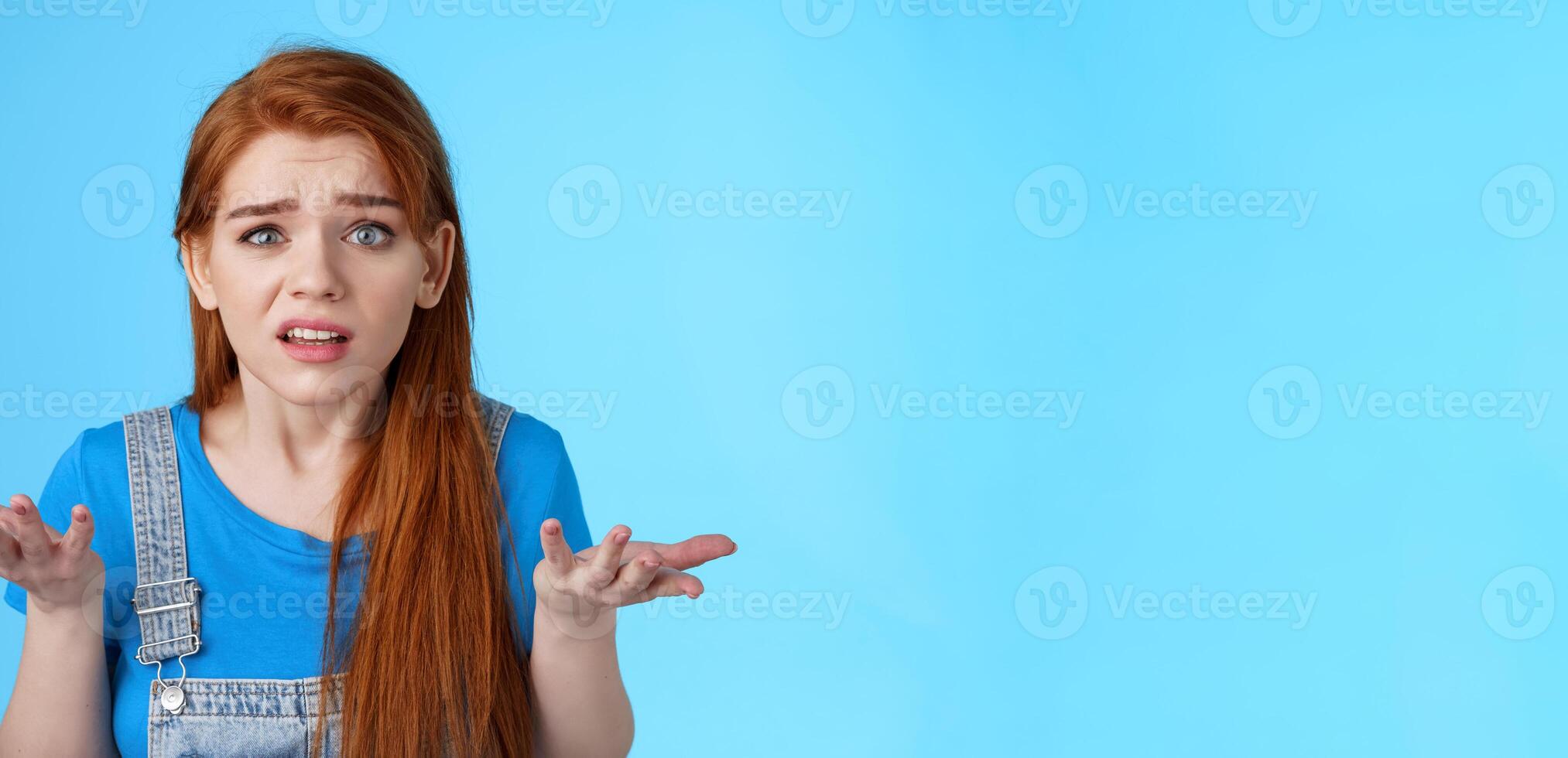Puzzled confused redhead cute woman look ambushed, raise hands shrugging uncertain, frowning perplexed, cannot understand what happened, stand frustrated upset blue background photo