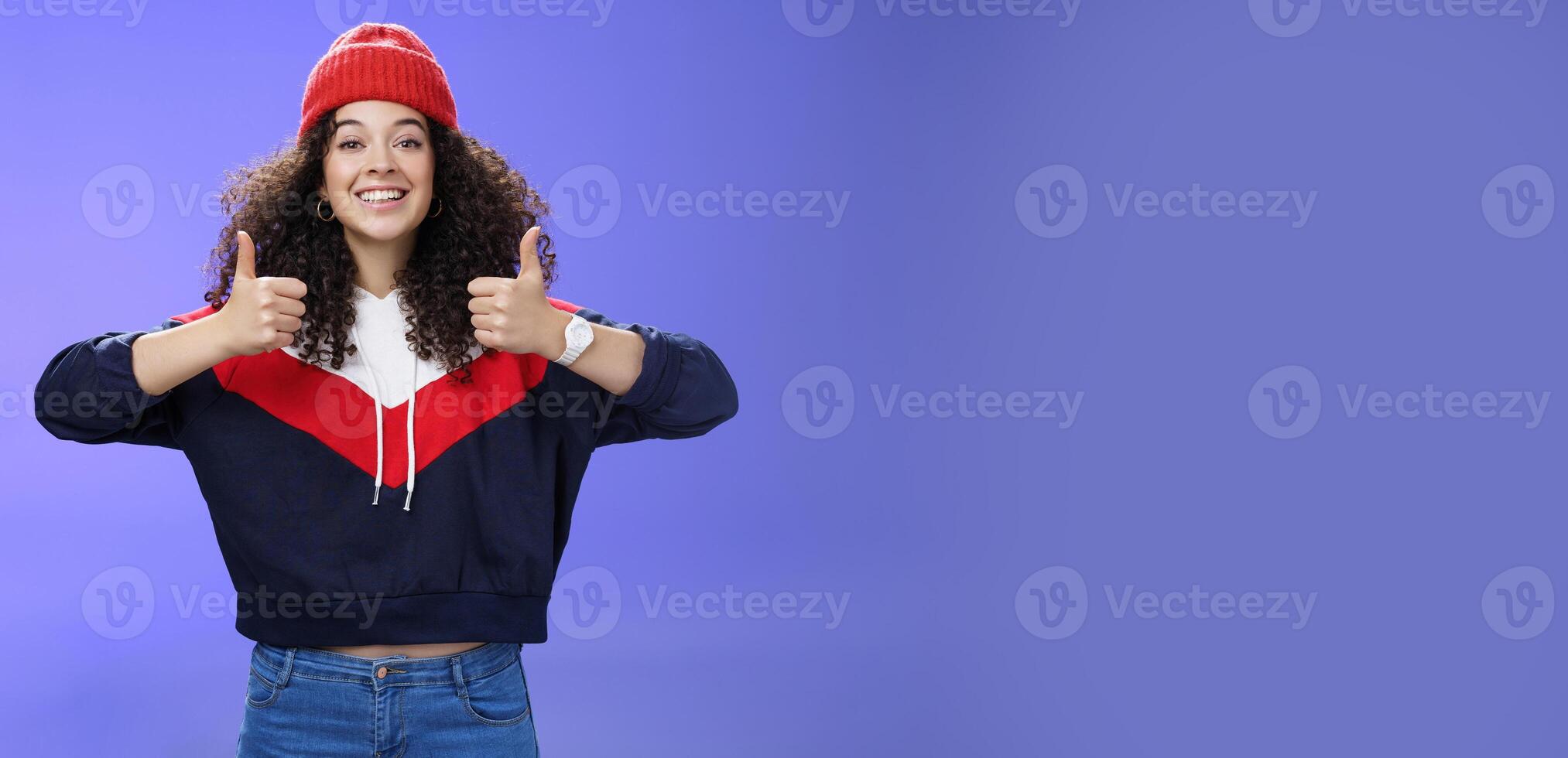 Girl totally agrees recommends cool place showing thumbs-up and smiling excited with happy grin standing upbeat and delighted against blue background in warm beanie and sweatshirt photo