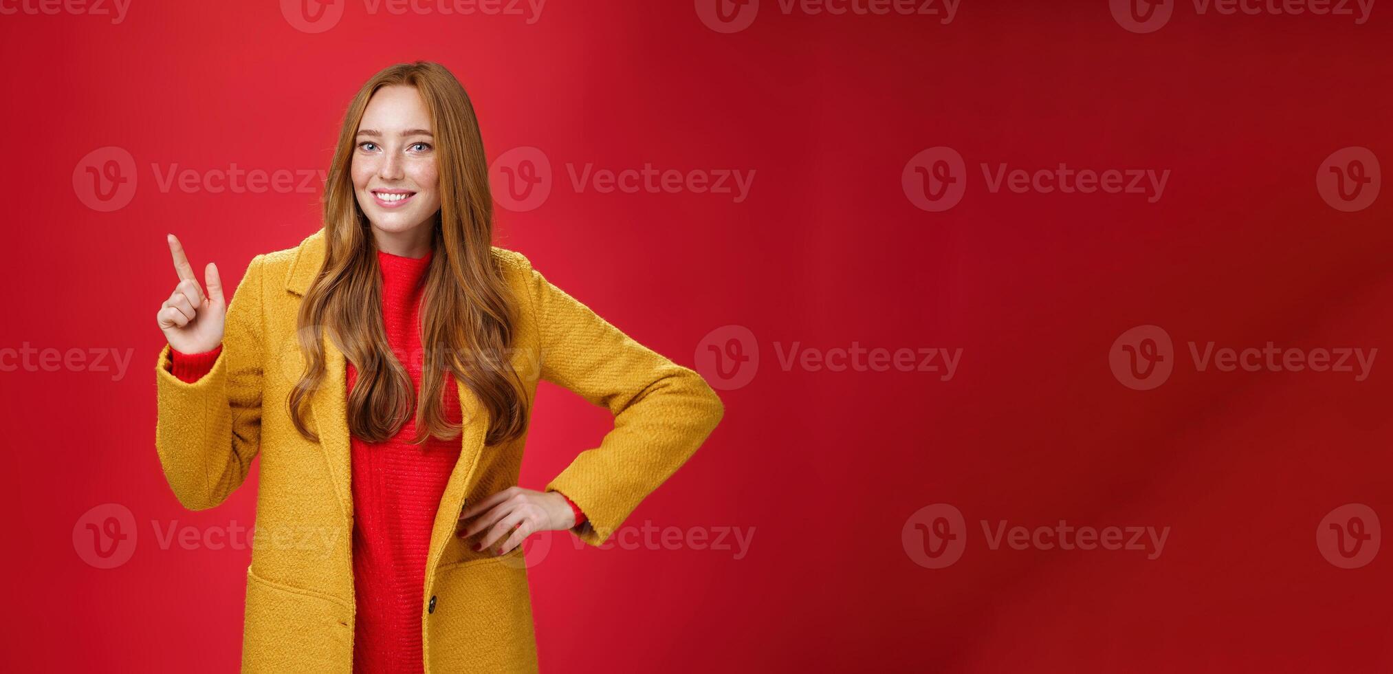 Charming young female in yellow coat holding hand on waist and pointing at upper left corner, smiling broadly as showing or promoting interesting proposal over red background, in good mood photo