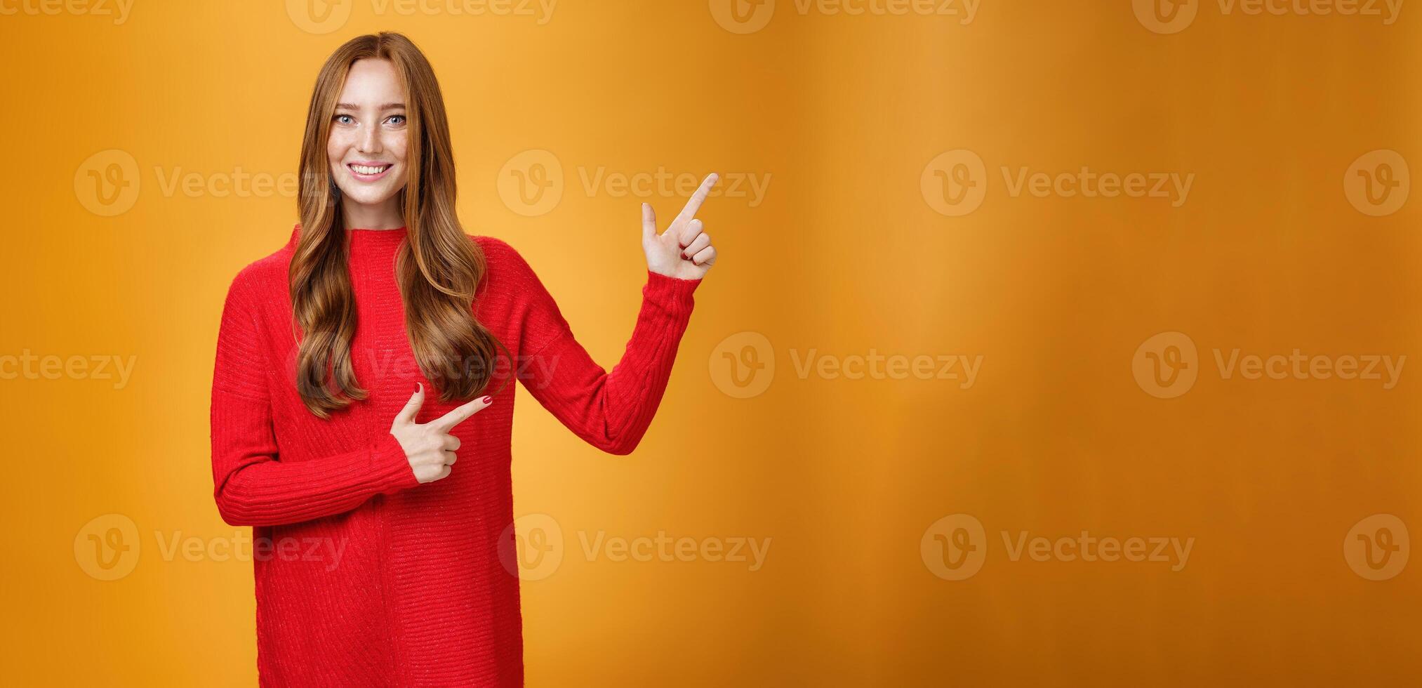 Friendly-looking joyfuly and energized redhead female in red sweater pointing at upper right corner promoting advertisement with broad delighted and tender smile posing against orange background photo