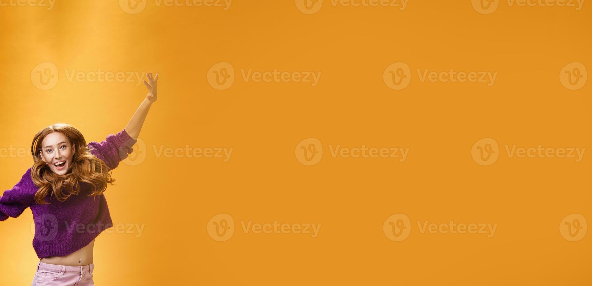 Expressive and enthusiastic carefree friendly joyful redhead woman in purple sweater and pants jumping joyfully with raised hands smiling yelling from happiness and joy against orange background photo