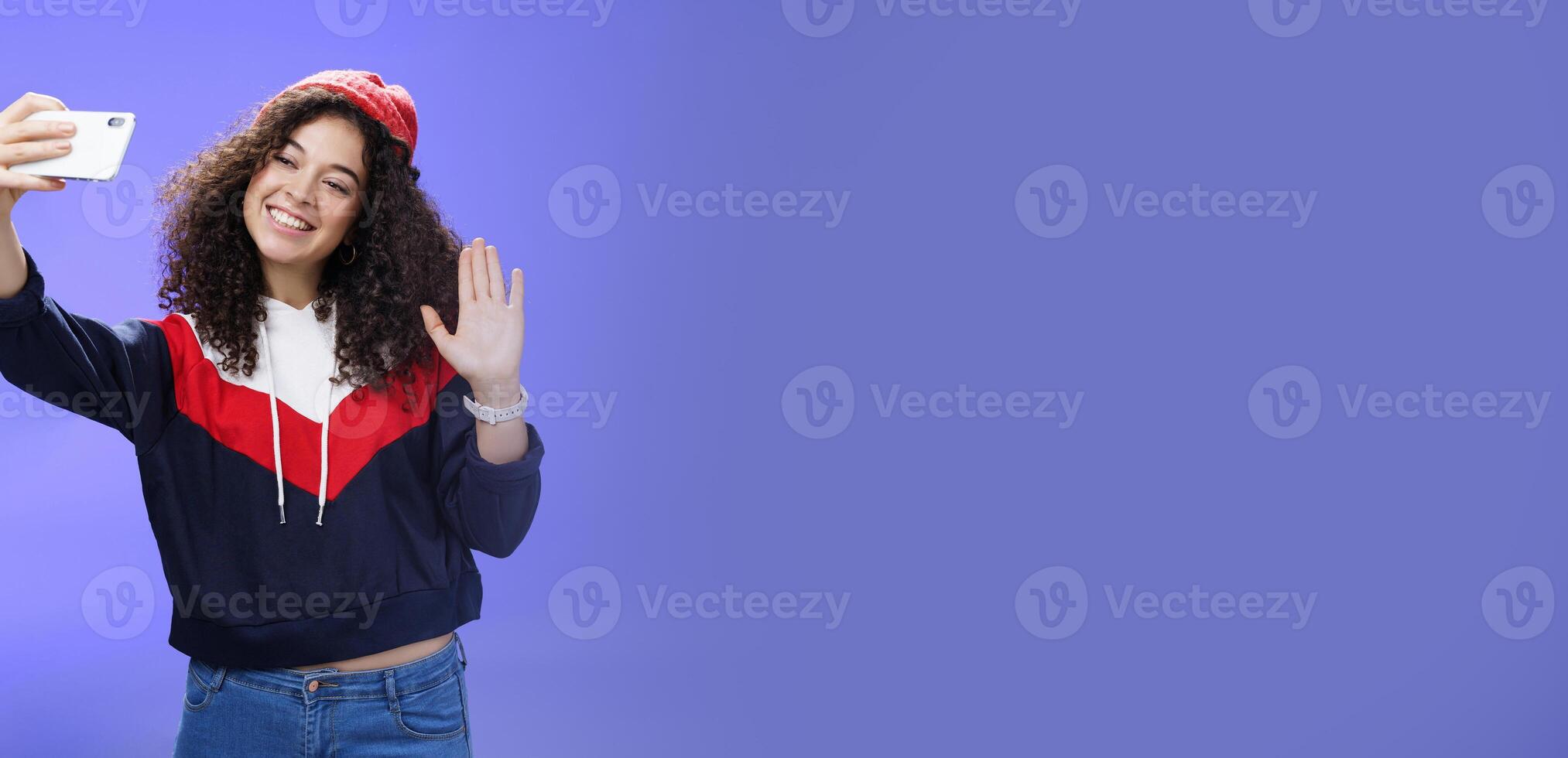 Studio shto of stylish cute female vlogger with curly hair in winter red hat smiling and waving hello at smartphone camera as recording , making interesting content to post online over blue wall photo