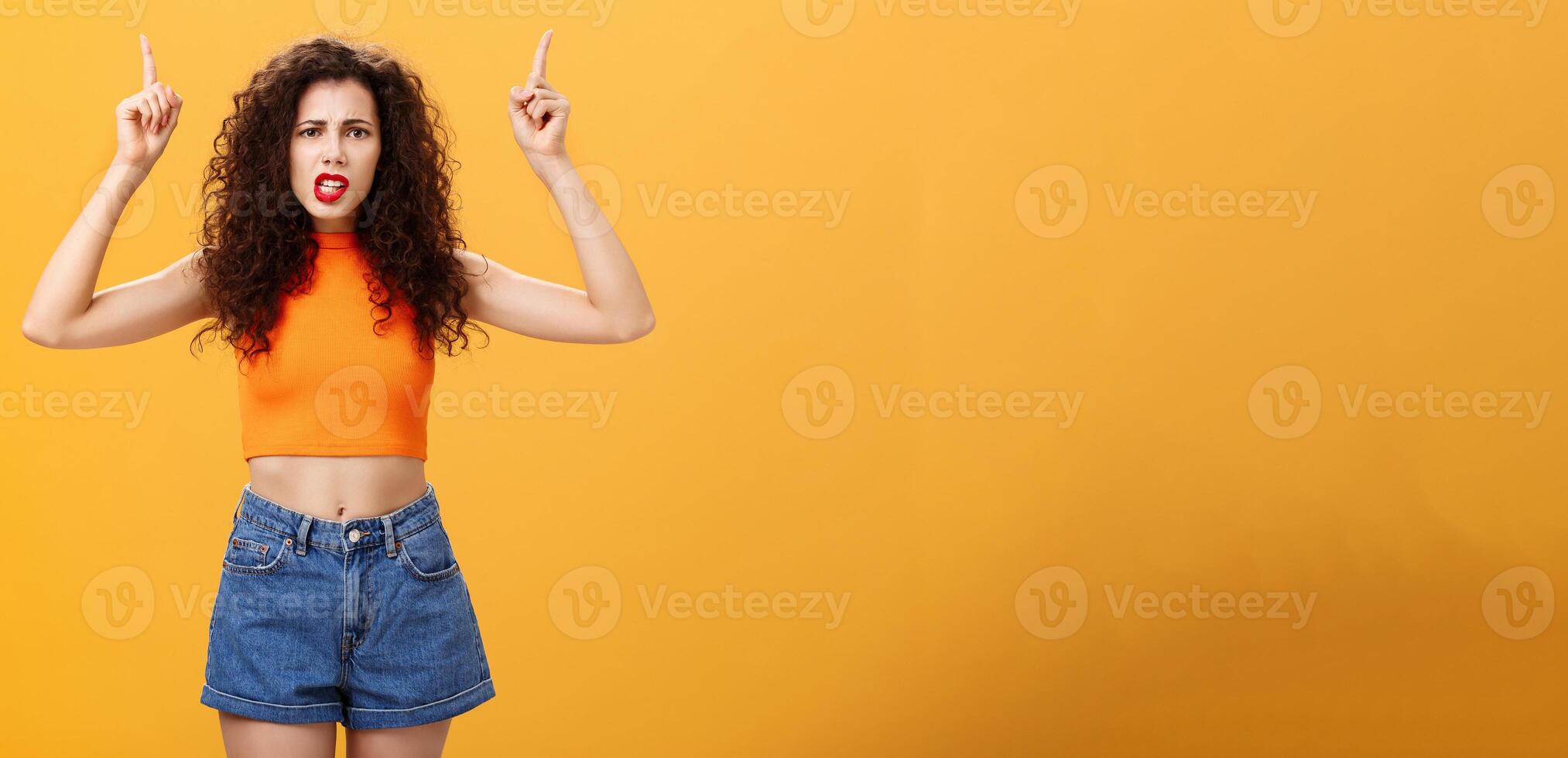 Indoor shot of displeased female client with curly stylish hairstyle red lipstick in cool cropped top pointing up demanding explanation with confused and dissatisfied expression over orange background photo