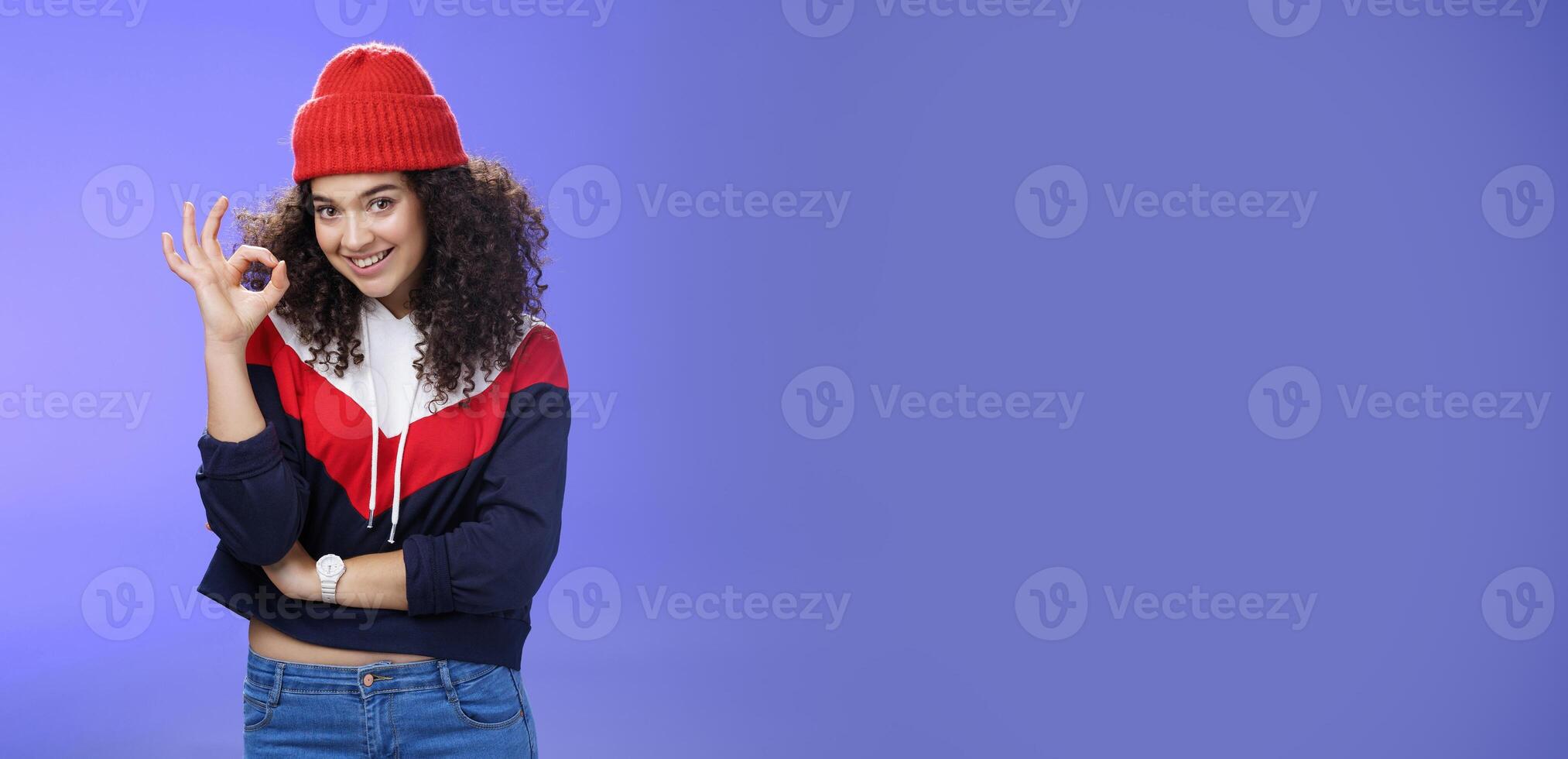 Portrait of pleased good-looking assertive woman with confident look and curly hair wearing warm winter hat showing okay gesture and smiling assuring, liking and approving awesome outfit of friend photo