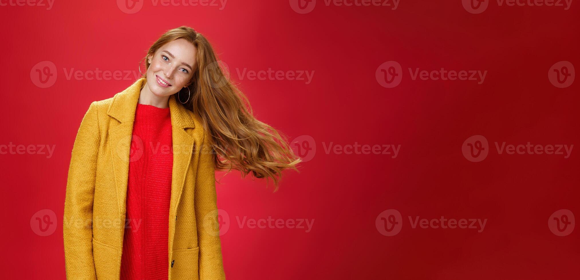 Charming redhead female with freckles waving hair smiling broadly tilting head as haircut flying in air posing delighted and carefree over red background in warm yellow coat and knitted dress photo