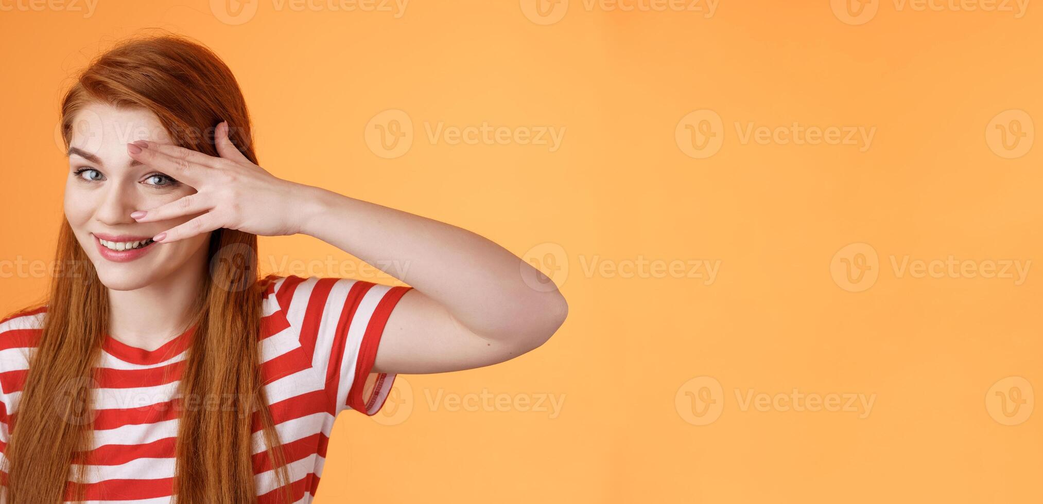 Charismatic lovely redhead lucky female, smiling cute, peek camera tender coquettish gaze, hold palm face, look through fingers, stand orange background optimistic, cheerful mood photo