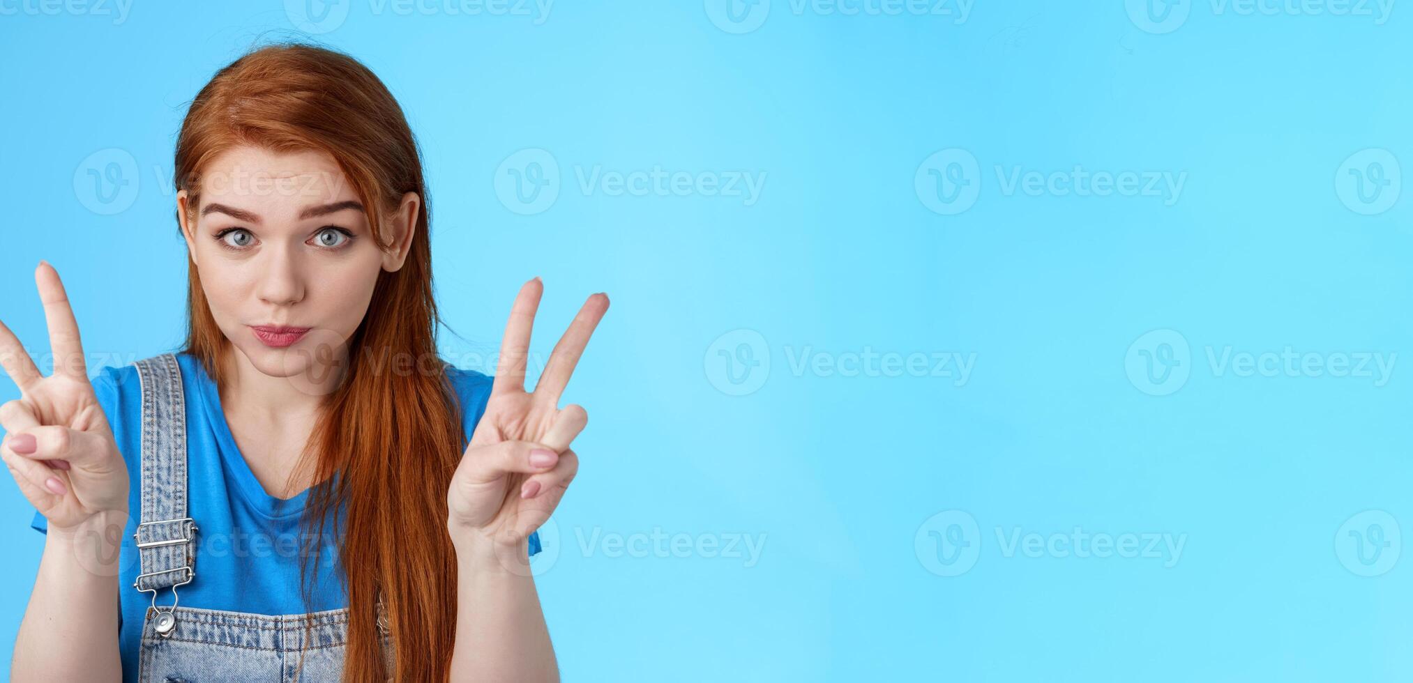 Playful cute silly redhead girlfriend making funny face, show peace victory signs, hold breath, pouting childish, fool around having fun, stand blue background, relaxing entertain siblings photo