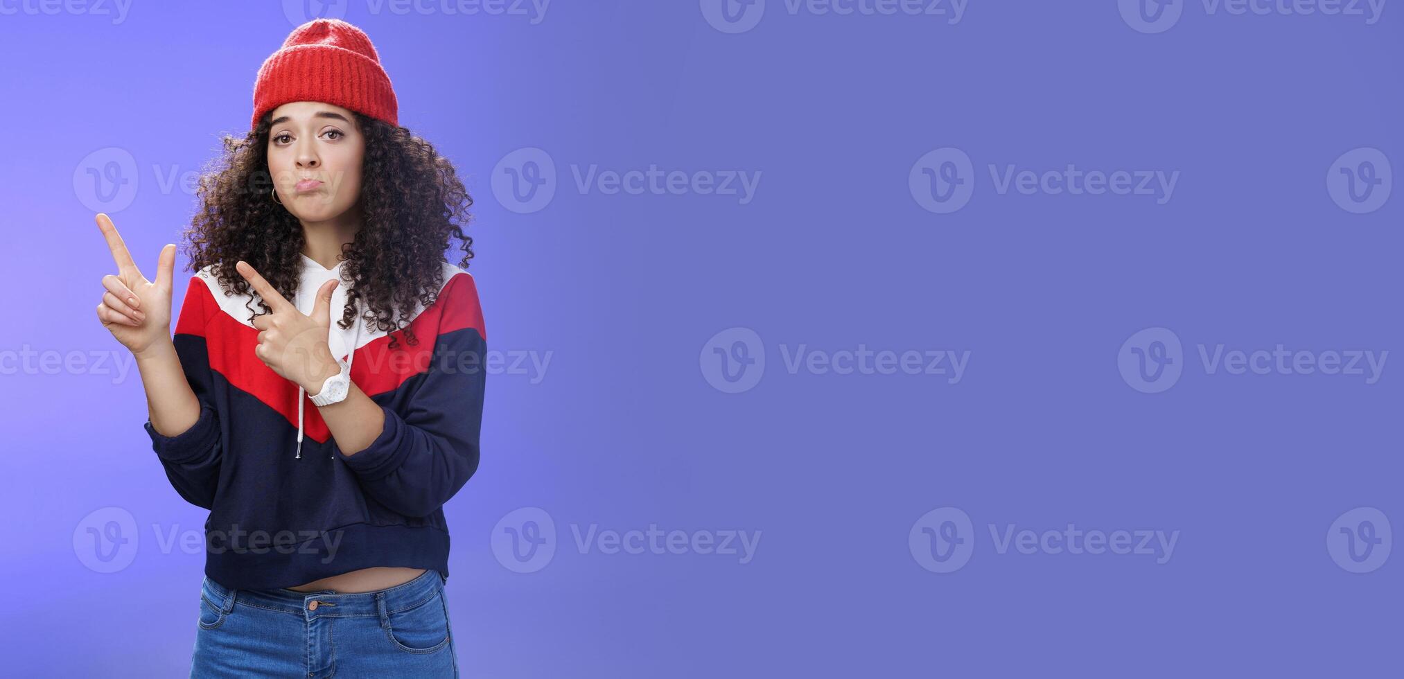 Portrait of silly upset cute curly-haired woman in trendy beanie pursing lips in sadness and frowning making disappointed, regret face pointing at upper left corner unhappy and sad over blue wall photo