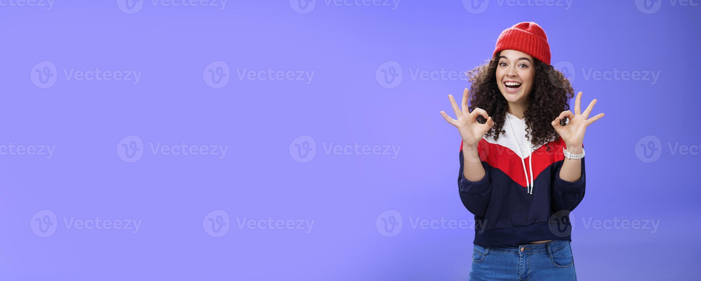 Got under control. Portrait of happy charming smiling curly-haired female in warm winter hat and sweatshirt smiling broadly and showing okay or excellent gesture as approving, liking cool movements photo