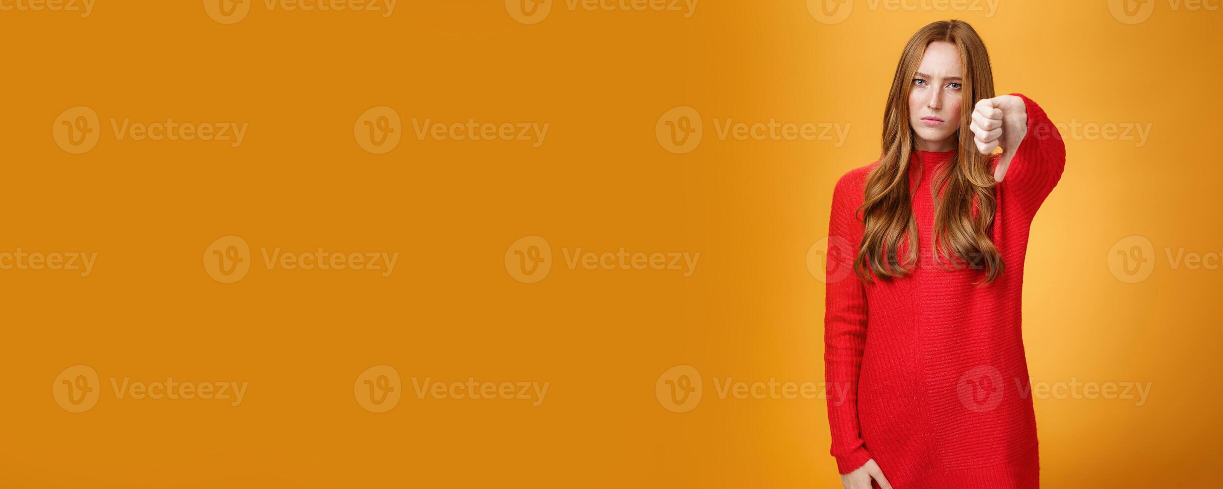 Disappointed upset and serious-looking attractive 20s redhead woman in red knitted dress frowning in dislike looking with judgement, showing thumb down in aversion and dissatisfaction over orange wall photo