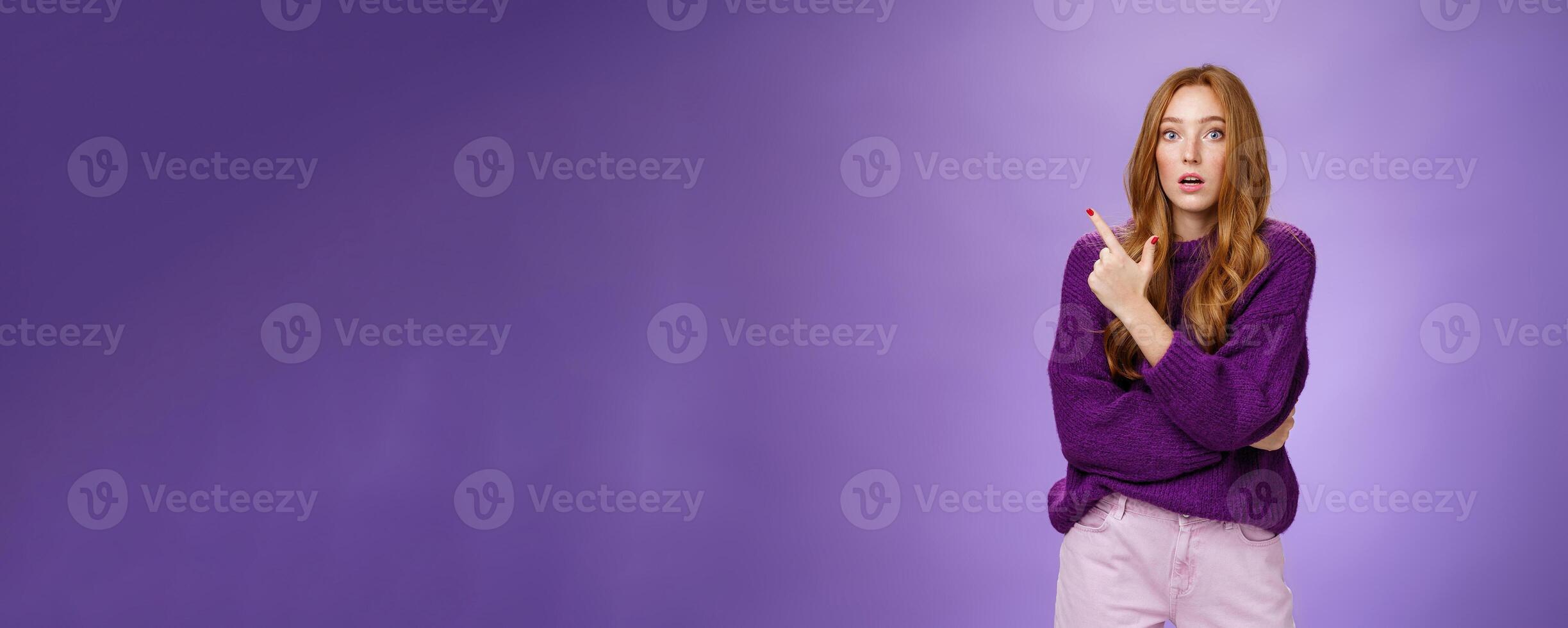 Astonished excited redhead woman open mouth and raise eyebrows questioned as seing shocking thing pointing at upper left corner, asking question impressed and surprised over purple wall photo
