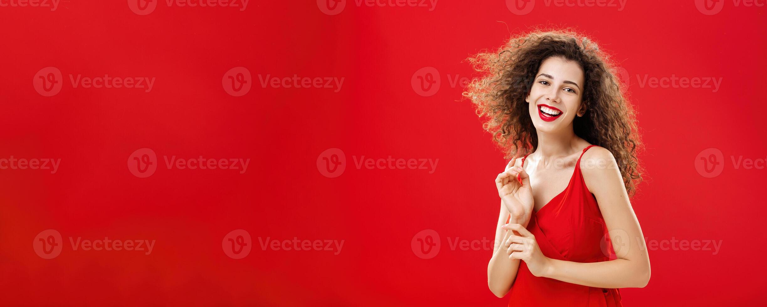 Waist-up shot of tender and elegant carefree happy woman with curly hairstyle in red stylish evening dress smiling cute and delighted while hair flick on air standing timid and gentle photo