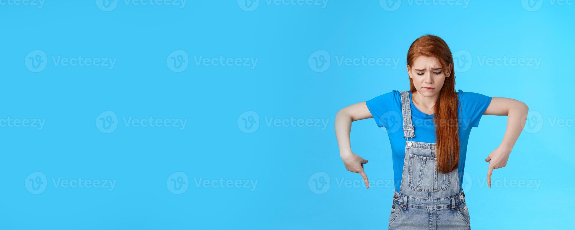 Moody upset cute redhead woman feel uneasy, frowning look pointing up jealousy and regret, cannot afford awesome new product, express sadness and sorrow, stand blue background gloomy photo