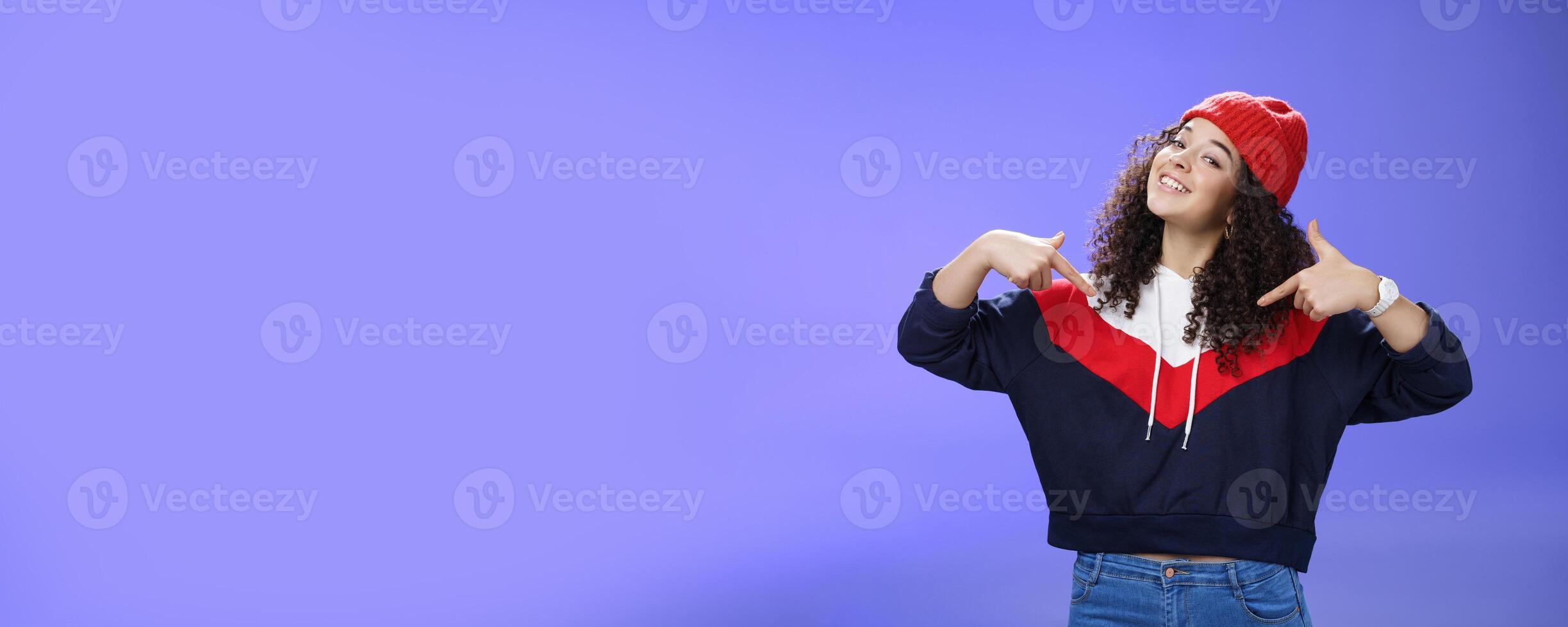 Proud and self-satisfied happy young energized stylish female model in warm beanie brag about own achievements as pointing at herself raising head and smiling produly posing delighted over blue wall photo