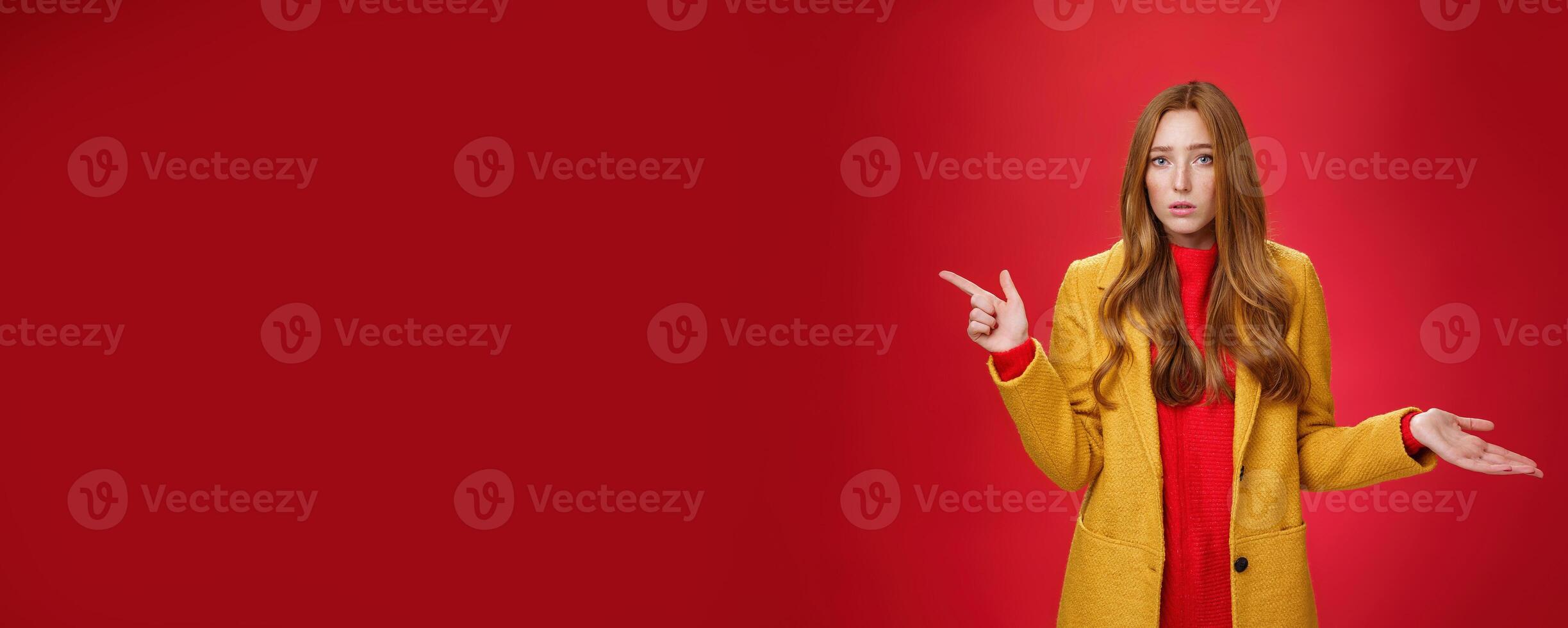 Frustrated and confused young cute female with ginger hair in yellow coat shrugging as pointing questioned left waiting explanation as asking question clueless and puzzled over red background photo