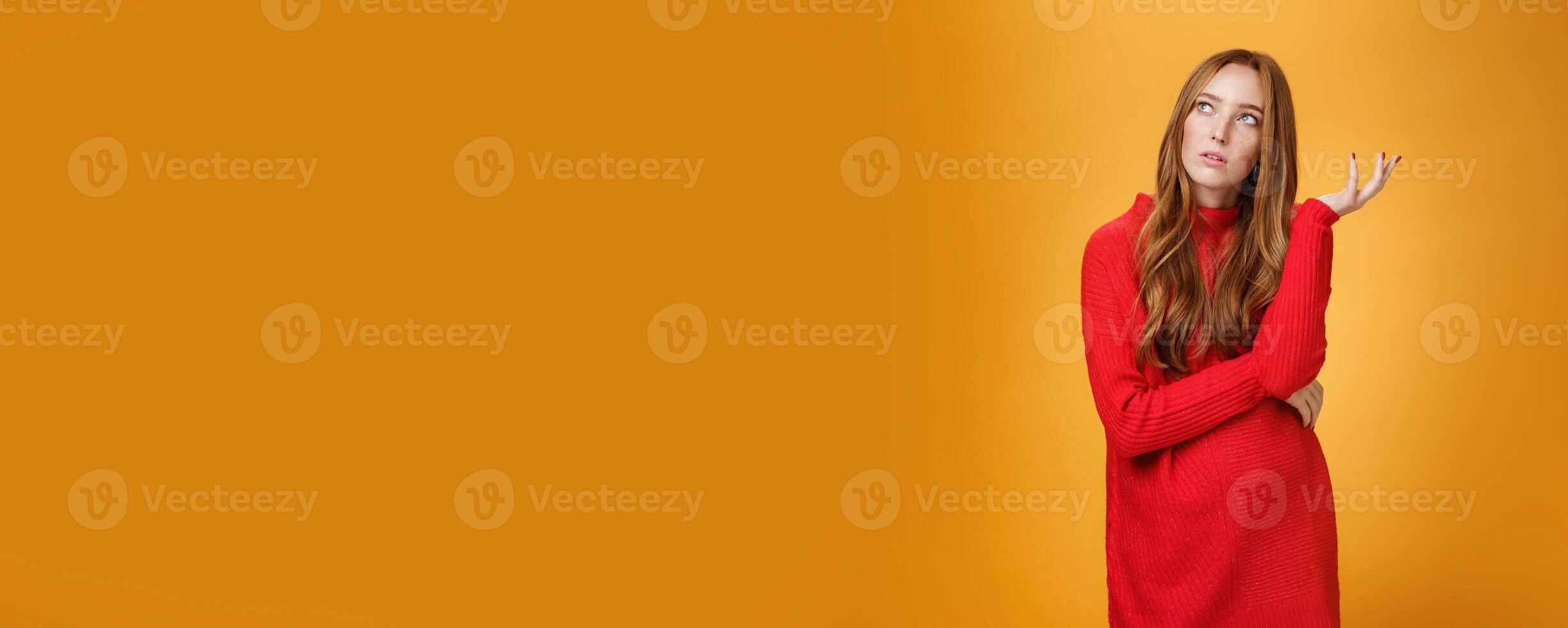 Portrait of unsure thinking redhead girlfriend in red sweater looking uncertain at upper left corner, recalling, feeling nostalgic, solving puzzle in mind, posing against orange background photo
