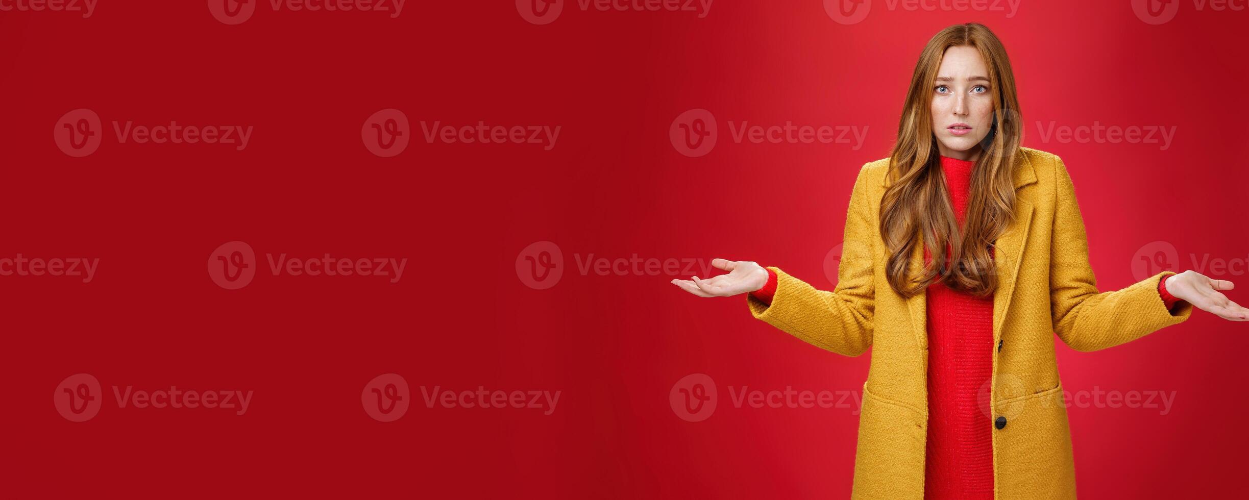 Upset and confused redhead girlfriend cannot understand what happened shrugging with spread hands sideways and clueless grimace, being unaware why, posing concerned over red wall photo