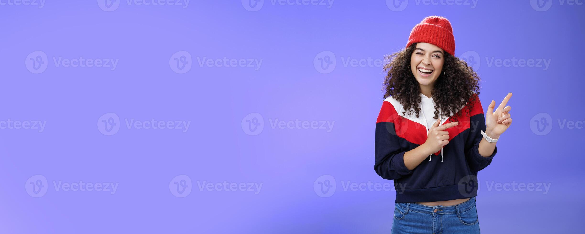 Charismatic playful young curly-haired european female winking joyfully at camera as pointing at upper right corner wearing hat and pullover as posing delighted over blue background, feeling warm photo