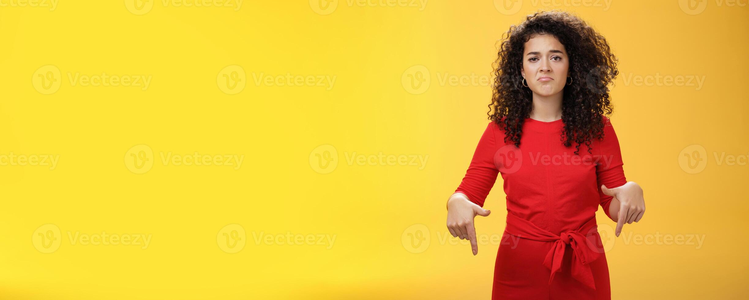 Portrait of gloomy girl with sad face pursing lips down frowning disappointed and pointing down with regret and unsatisfied expression, missing opportunity feeling upset over yellow background photo