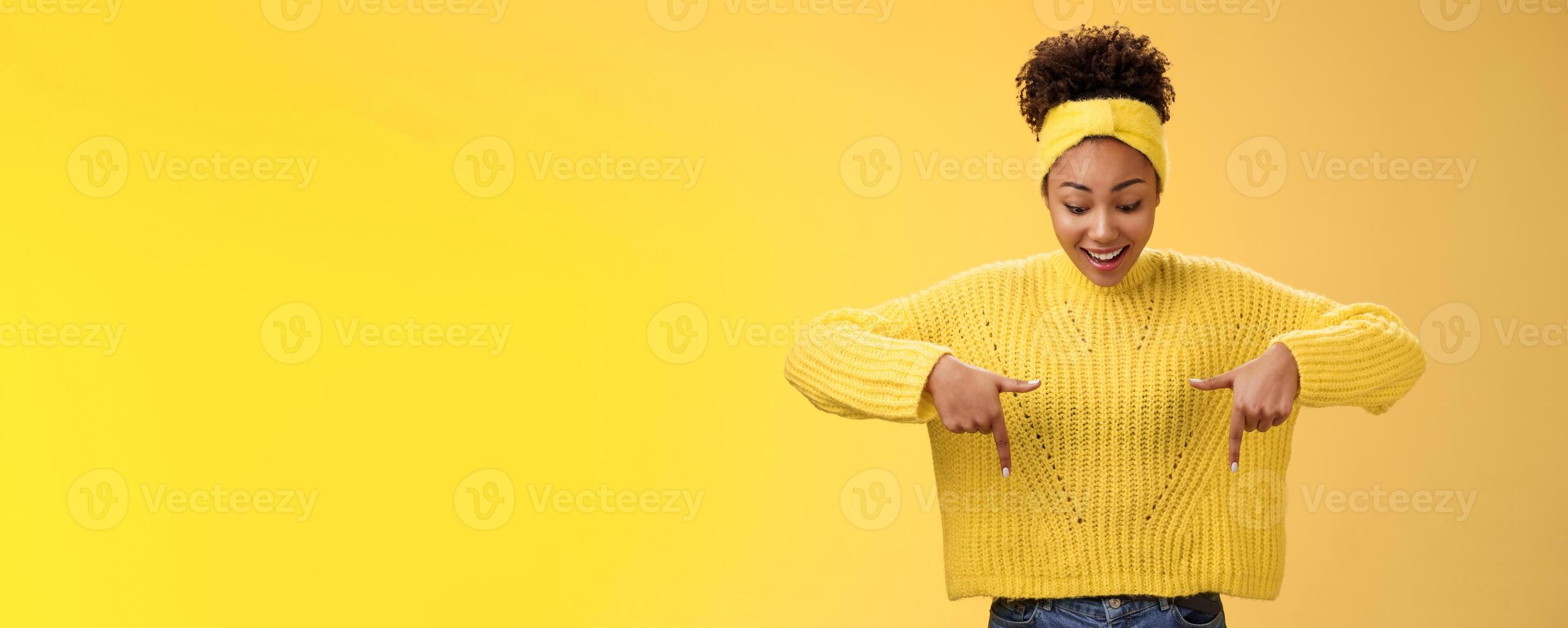 Impressed enthusiastic smiling fascianted african-american woman in sweater headband look pointing down index fingers gasping astonished fascinated cool thrilling promo, standing yellow background photo