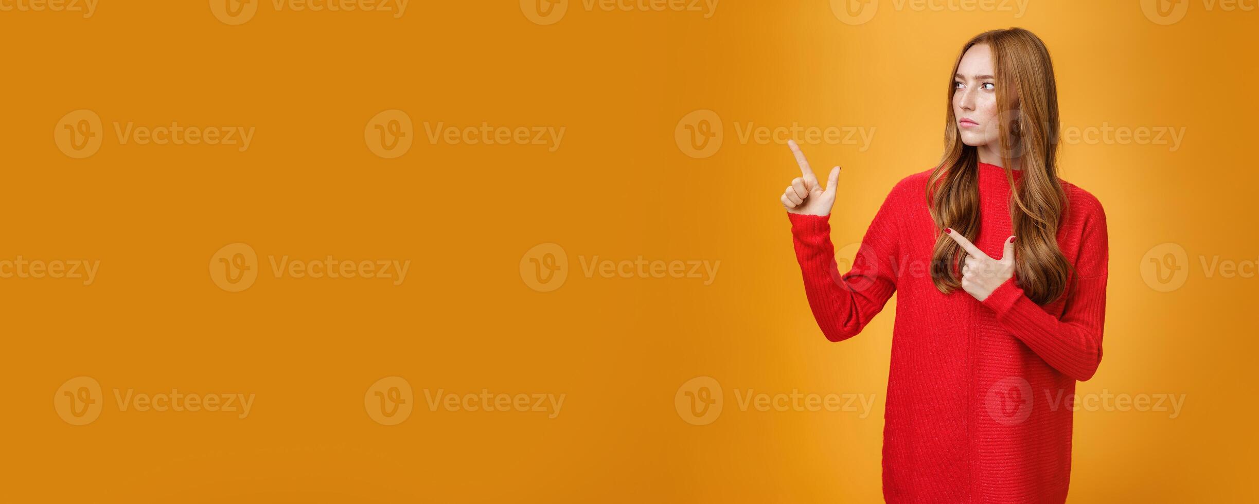 Studio shot of suspicious serious-looking unsure redhead girl frowning looking and pointing at upper left corner with doubtful strict gaze posing uncertain over orange background photo