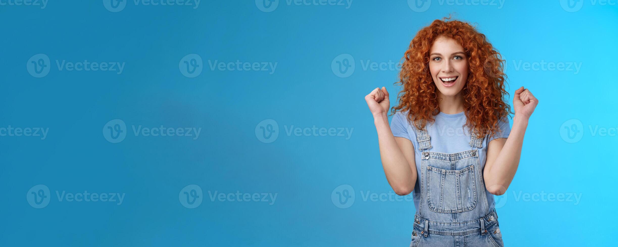 Hooray successful day. Cheerful chrismastic redhead girl summer dungarees clench fists joyfully smiling broadly determined achieve success, triumphing winning game standing upbeat blue background photo