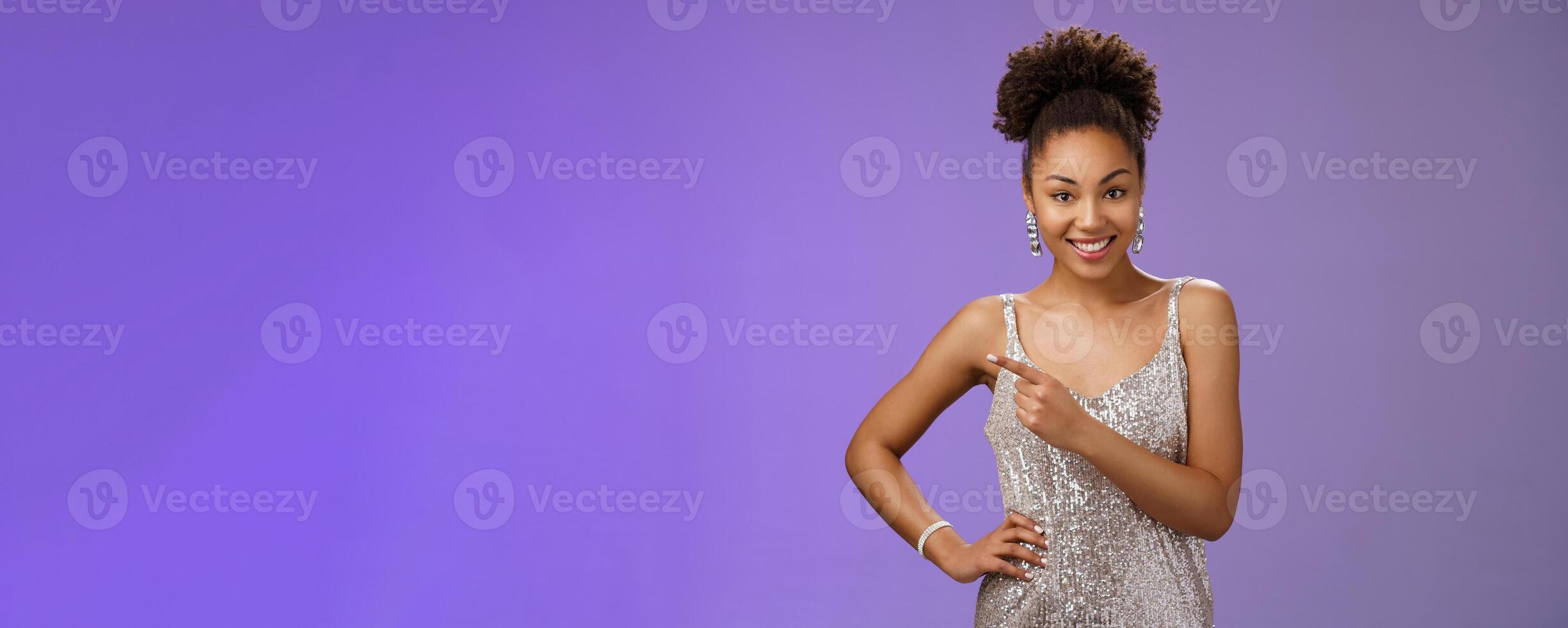 Elegant good-looking young b-day girl partying in silver stylish glittering dress pointing left index finger smiling delighted friendly chat showing place guests standing blue background sociable photo