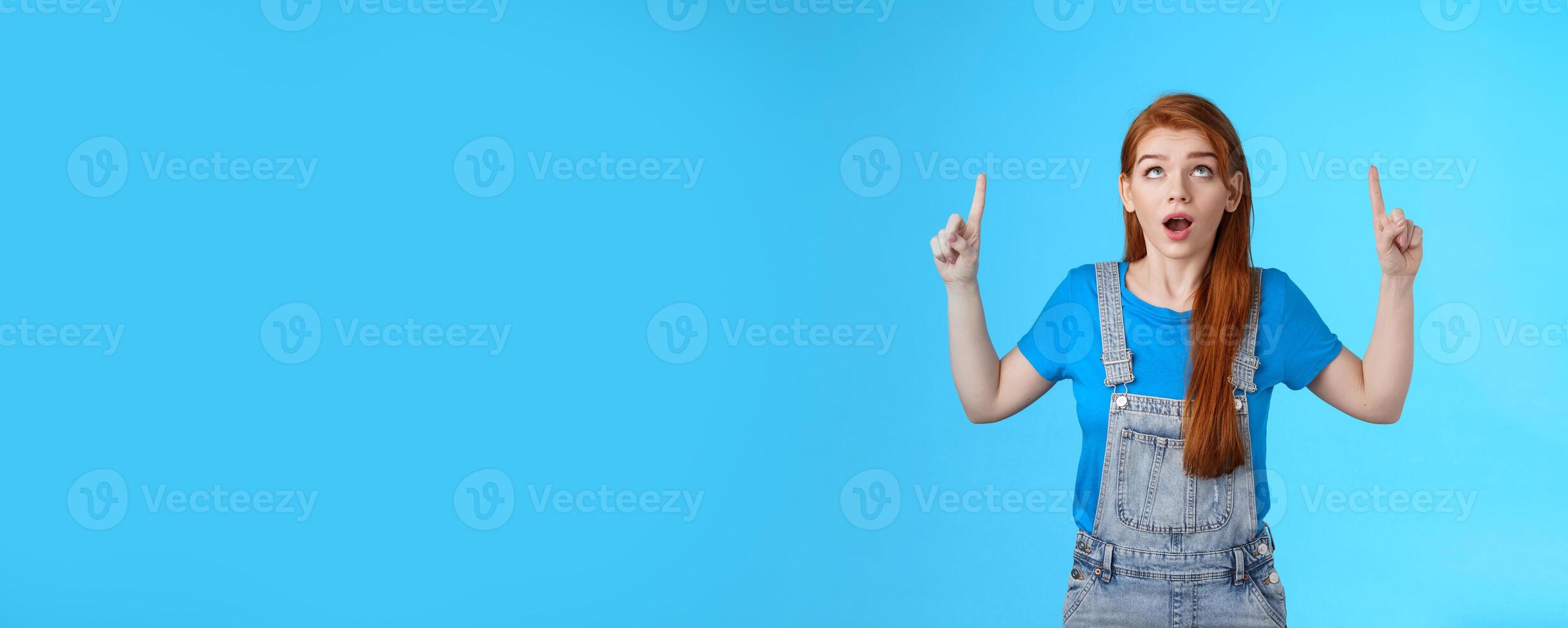 Studio shot amazed curious redhead woman drop jaw astonished, look pointing ambushed top copy space, check out up advertisement thrilled, stand surprised blue background open mouth photo