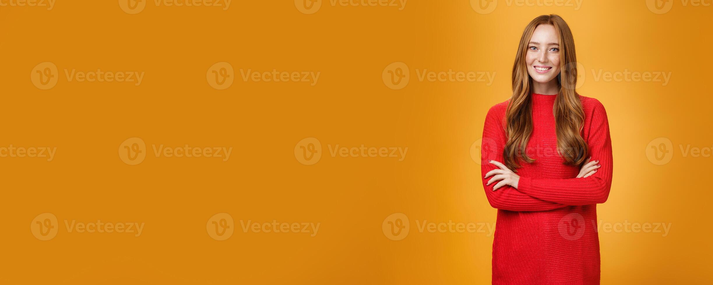 Portrait of elegant and friendly, polite redhead female in red knitted dress holding hands crossed over body in confident and relaxed pose smiling satisfied offering help over orange background photo