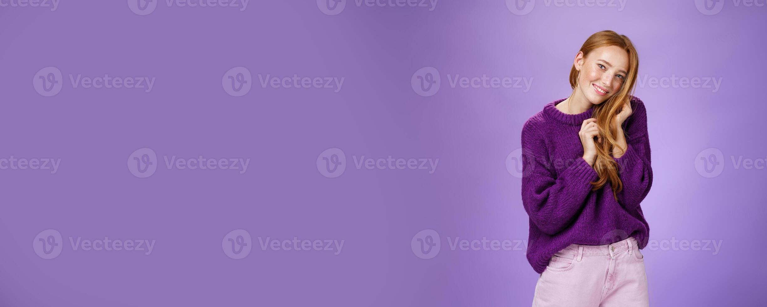 Tender and flirty feminine redhead woman playing with hair tilting head to shoulder and smiling cute and sensual as if making gazes of admiration, being coquettish over purple background photo