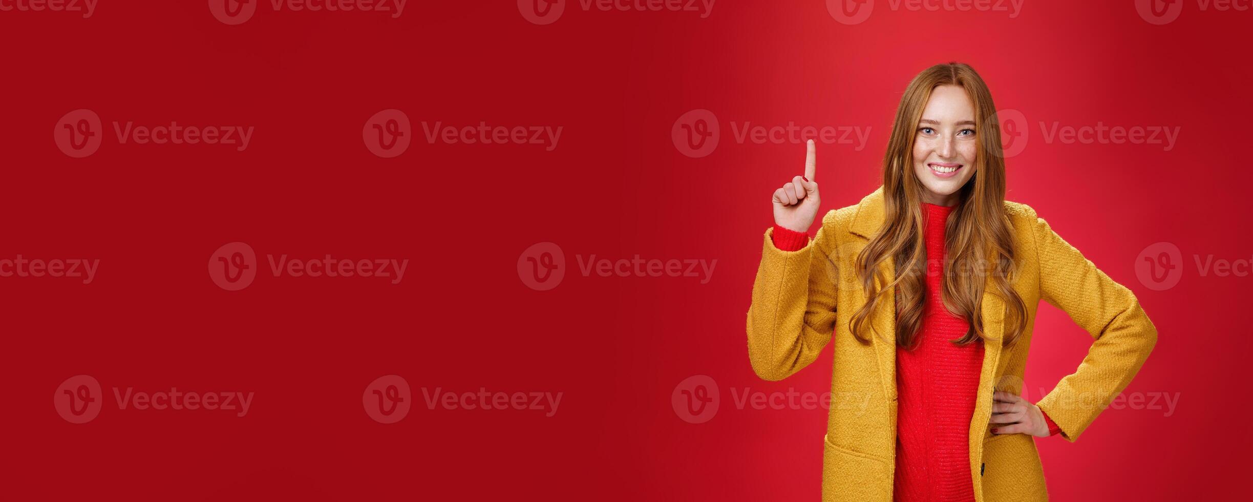 Girl telling first reason use her advice. Cute and friendly energized young redhead woman in yellow coat raising hand showing number one and smiling broadly at camera against red background photo