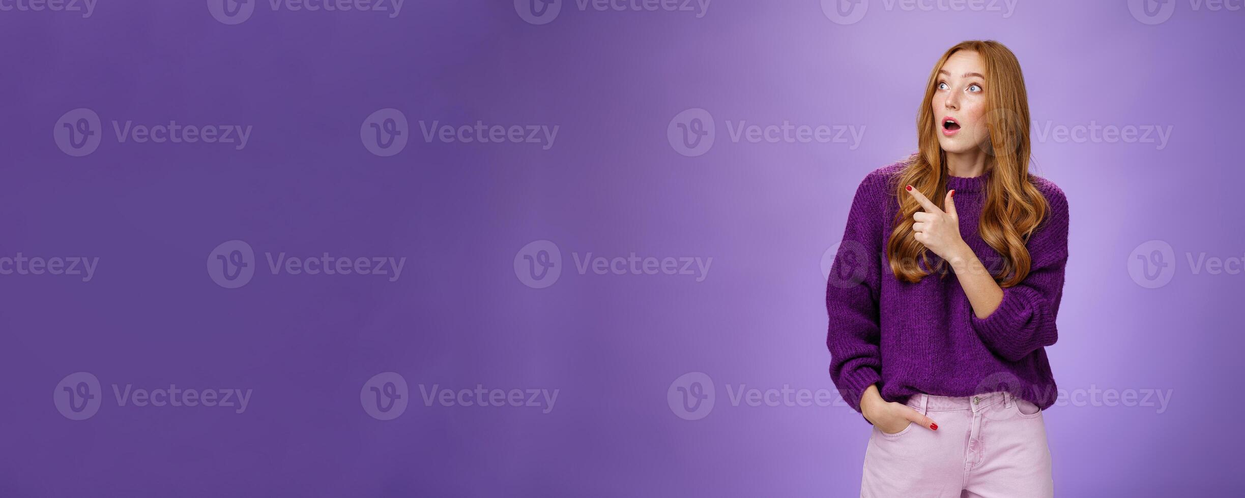 Girl surprised and thrilled as seing unbelievable scene. Portrait of impressed and shocked good-looking ginger woman in purple sweater gasping from amazement pointing, looking left intrigued photo